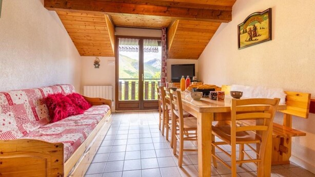 Una stanza accogliente con soffitto in legno, un divano con una coperta rossa, un tavolo da pranzo con sedie e vista all'esterno tramite una porta.