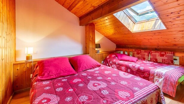 Una camera accogliente con soffitto spiovente e finestra sul tetto, con due letti con biancheria rossa coordinata e un comodino con lampada.