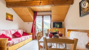 Una camera accogliente con soffitto in legno, un divano con cuscini rossi, un tavolo da pranzo con sedie, una TV a parete e vista sulle montagne dalla finestra.