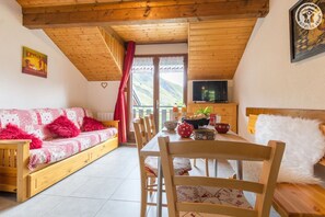 A cozy room with a wooden ceiling, a sofa with red cushions, a dining table with chairs, a TV on the wall, and a view of the mountains through the window. - Les Balcons de St So Apartment B – 3 Bedrooms, Ski-In/Ski-Out, Sleeps 8 (Saint-Sorlin-d'Arves)