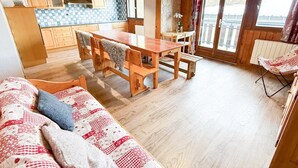 A cozy cabin interior with a wooden dining table, a sofa with patterned cushions, and a large window providing a view of the outdoors. - Les Balcons de St So Apartment D – Spacious, Sleeps 12, Near Slopes & Shops (Saint-Sorlin-d'Arves)