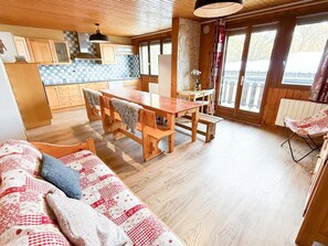 A cozy cabin interior with a wooden dining table, a sofa with patterned cushions, and a large window providing a view of the outdoors. - Les Balcons de St So Apartment D – Spacious, Sleeps 12, Near Slopes & Shops (Saint-Sorlin-d'Arves)