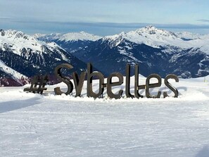 Ein Metallschild mit dem Text '#Sybelles' vor einer gebirgigen Landschaft.