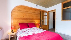A bedroom with a wooden headboard, a bed with red bedding, two bedside lamps, and a door leading to another room. - Les Balcons de St So Apartment 6 – 12 Guests, South-Facing Balcony, Near Slopes (Saint-Sorlin-d'Arves)