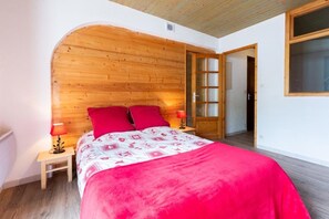 A bedroom with a wooden headboard, a bed with red bedding, two bedside lamps, and a door leading to another room. - Les Balcons de St So Apartment 6 – 12 Guests, South-Facing Balcony, Near Slopes (Saint-Sorlin-d'Arves)