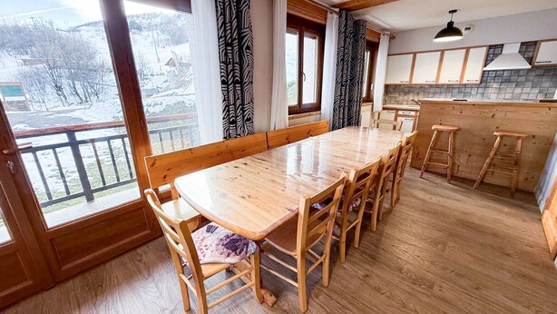 A wooden dining table with chairs, a kitchen area with cabinets, and a view of a snowy landscape through large windows. - Les Balcons de St So Apartment 6 – 12 Guests, South-Facing Balcony, Near Slopes (Saint-Sorlin-d'Arves)