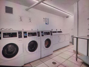 A laundry facility with multiple washing machines and dryers, a ventilation system, and a metal railing. - Cozy 1-bedroom 1.5 bath condo with AC in charming Hallandale Beach. Pool + gym. (Hallandale Beach)