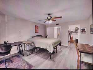 Quarto com cama, escrivaninha, cadeira, ventoinha de teto e guarda-roupa.