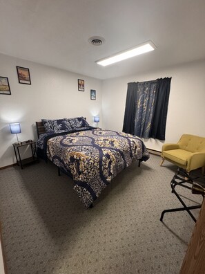 Une chambre d’hôtel avec un lit, une chaise, une table de chevet, une lampe et une fenêtre avec des rideaux.