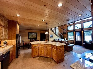 A spacious kitchen with a large island, wooden cabinets, and a dining area with a staircase leading to a loft. - Mack Mountain Lodge (Kirby)