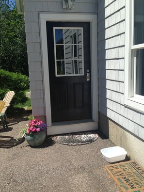 Une maison avec une porte d’entrée sombre, un petit porche, un pot de fleurs et un récipient blanc posé sur le sol.