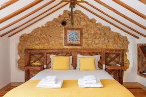 A bedroom with a stone headboard, wooden beams, a bed with white linens, and two yellow pillows. - GuestReady - Modern Duplex in Lisbon (Lisbon)