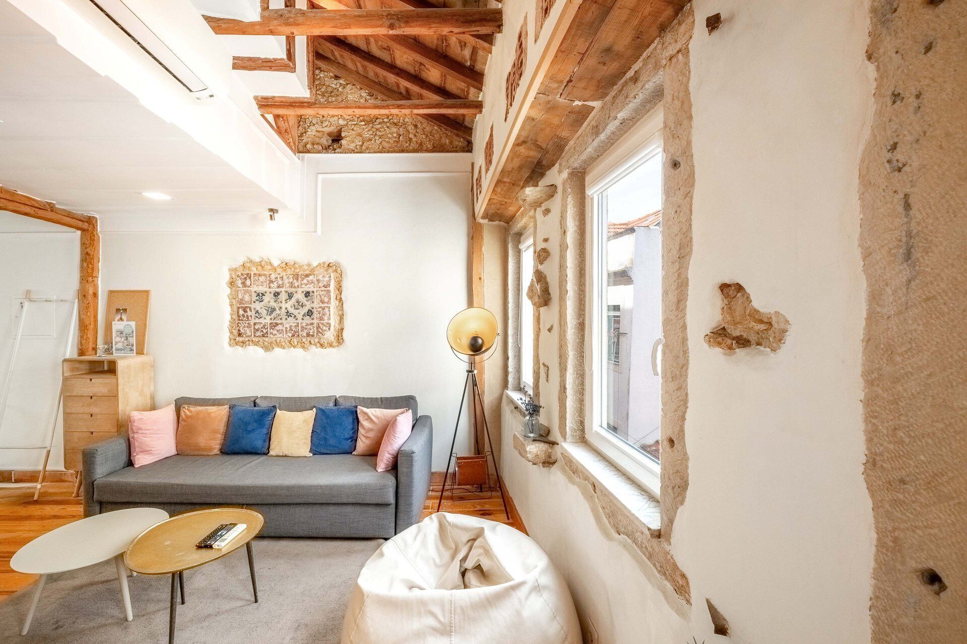 A cozy living room with a grey sofa, a coffee table, and a bean bag. There's a wooden ceiling with exposed beams and a window with peeling paint.