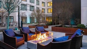 A fire pit surrounded by wicker furniture with cushions, set on a tiled patio with a building in the background.