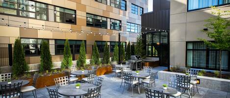 An outdoor patio with multiple tables and chairs, surrounded by a modern building with large windows and a water feature.