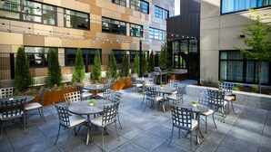 Un patio al aire libre con varias mesas y sillas, rodeado por un edificio moderno con amplios ventanales y una fuente de agua.