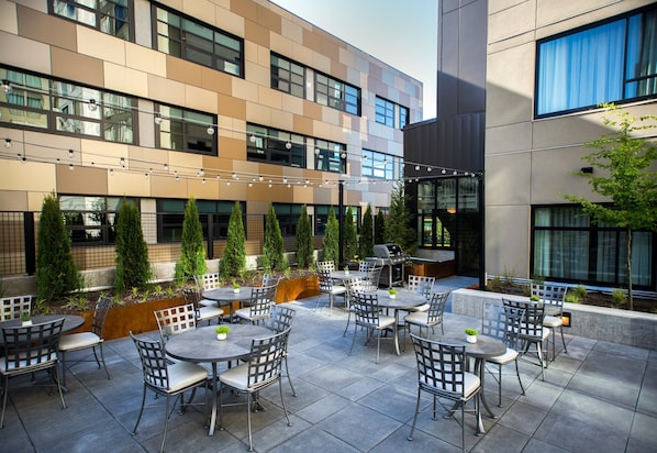Een modern terras buiten met meerdere tafels en stoelen, omringd door een gebouw met grote ramen en een waterpartij.