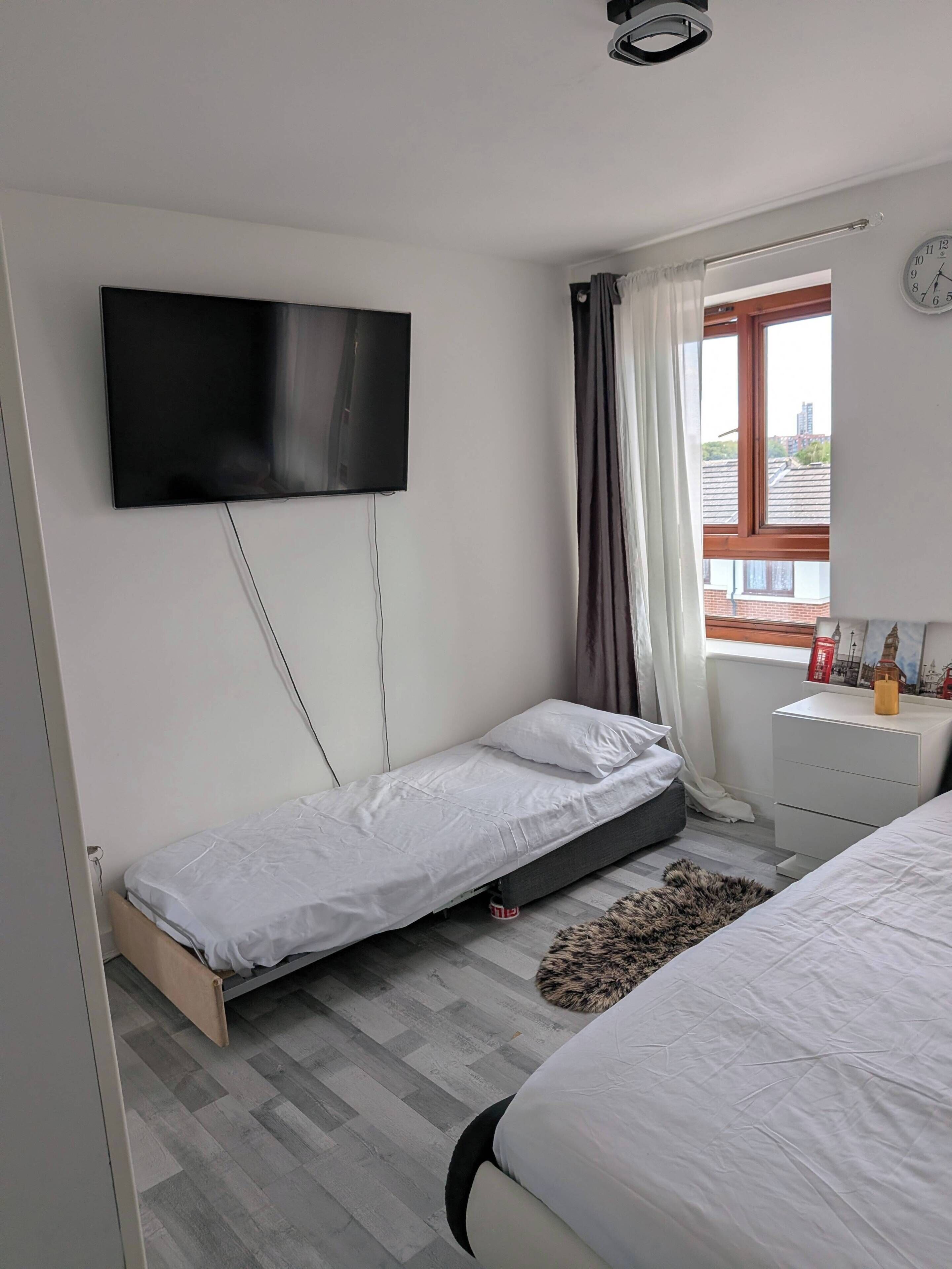 A bedroom with a single bed, a smaller bed, a TV mounted on the wall, a window with curtains, and a bedside table with a bottle.