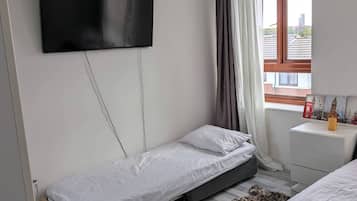 Une chambre avec un lit simple, un petit lit, un téléviseur fixé au mur, une fenêtre avec des rideaux et une table de chevet sur laquelle se trouve une bouteille.