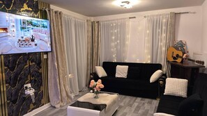A living room with a black sofa, white cushions, a coffee table with flowers, a television, and a guitar on a stand.