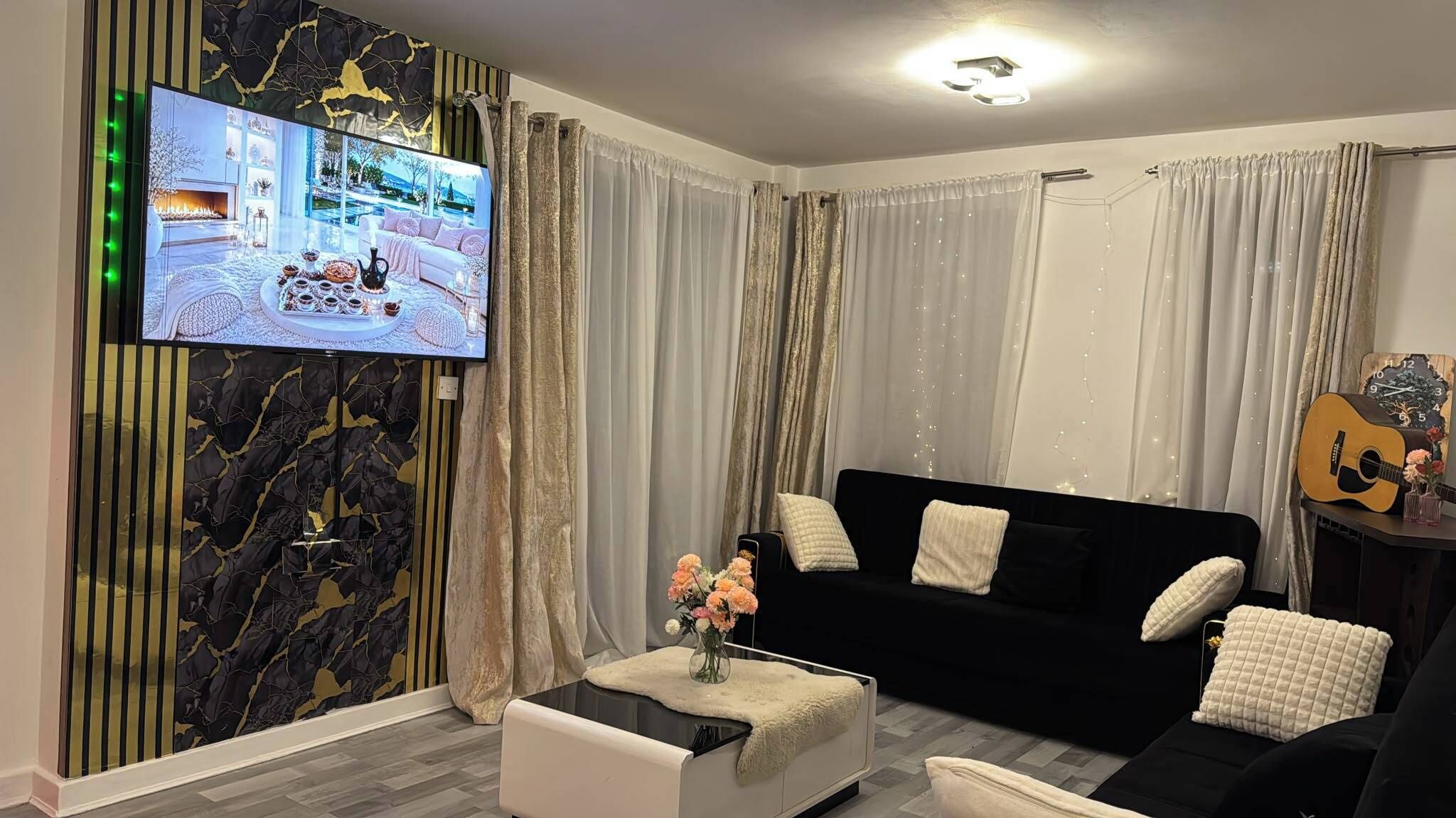 A modern living room with a black sofa, white cushions, a coffee table with a vase of flowers, and a television mounted on a wall with a marble-like pattern.
