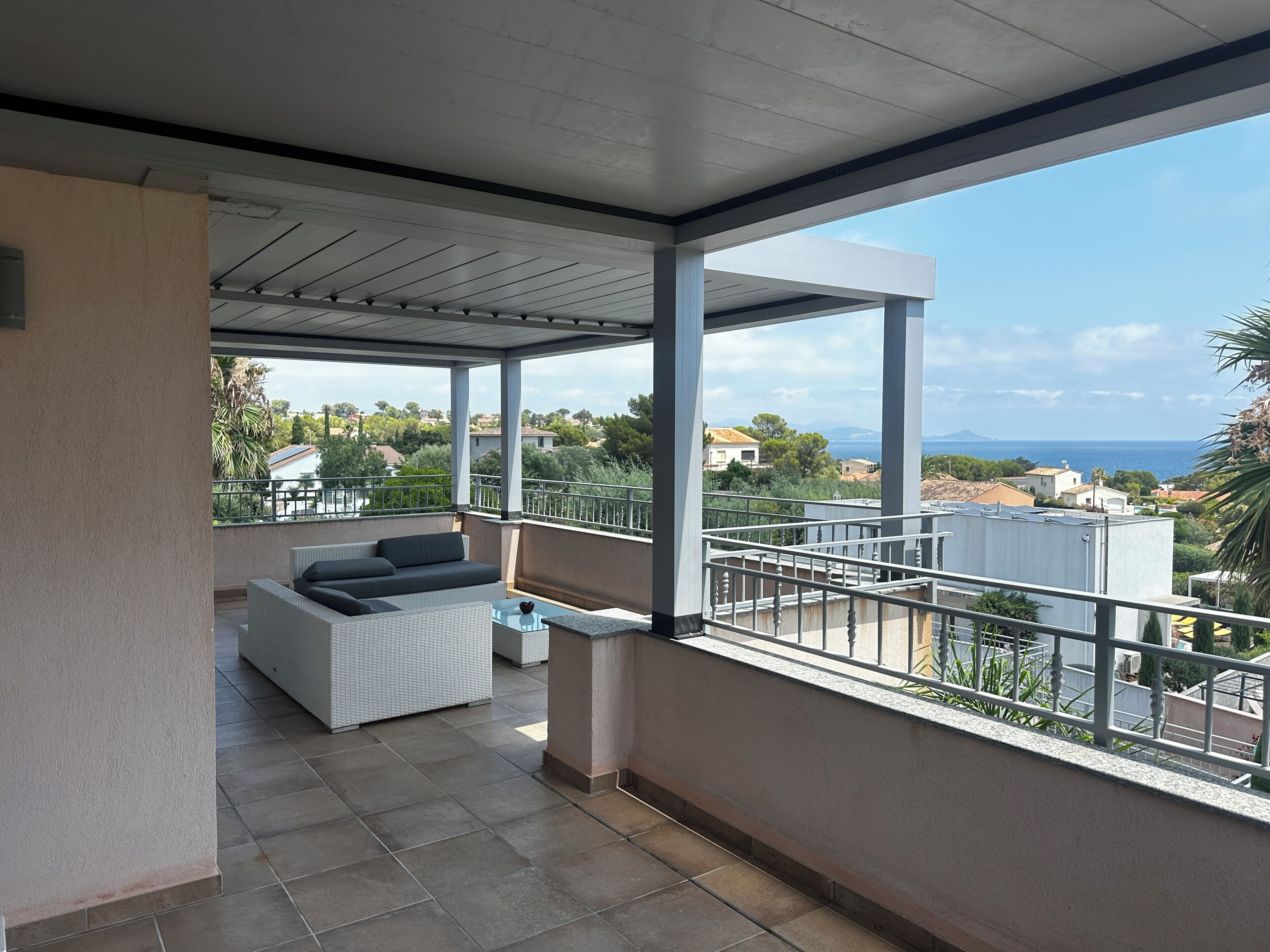 A covered outdoor balcony with a sofa and a small table, overlooking a residential area and the sea.