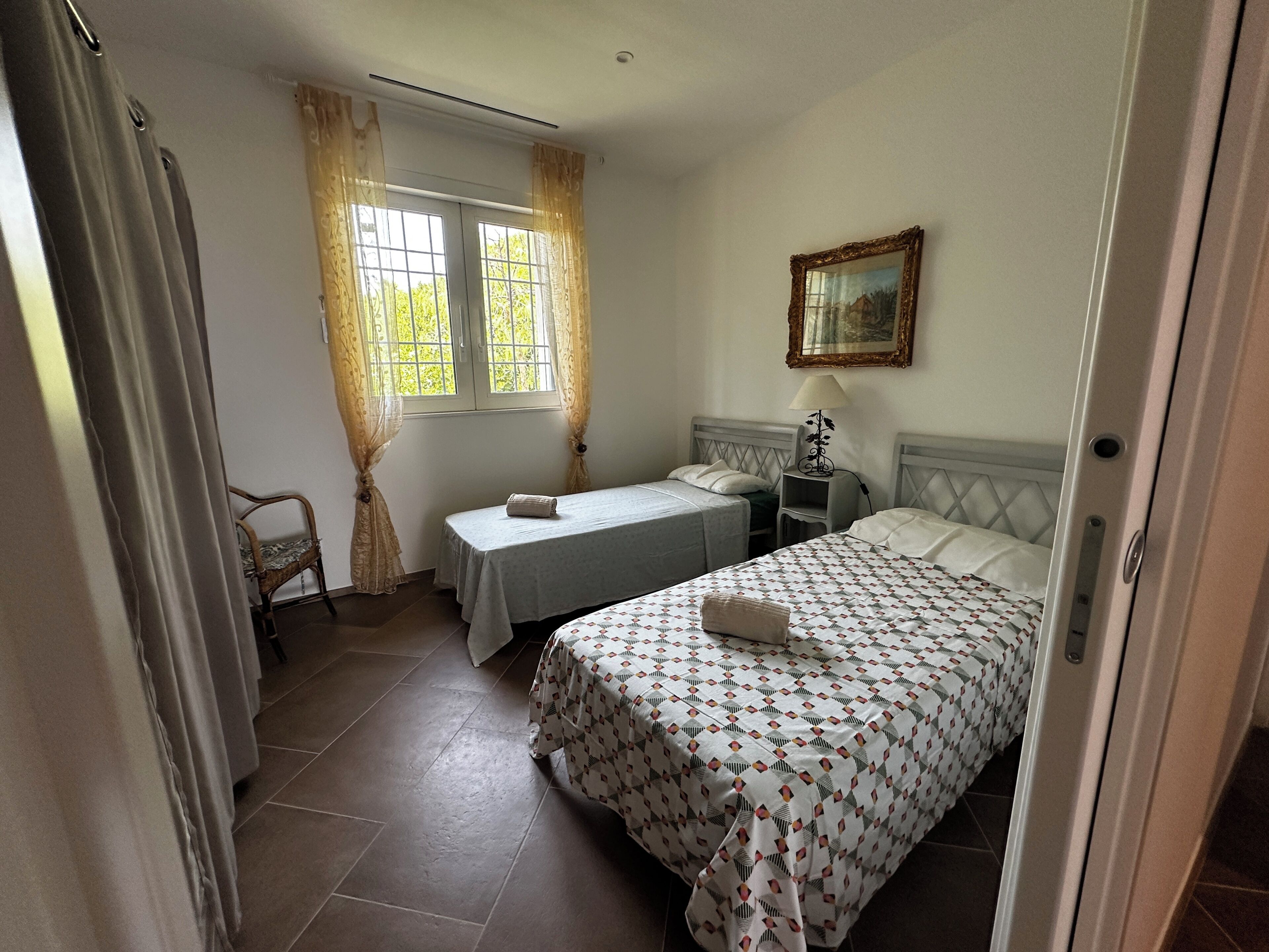 Ein Schlafzimmer mit zwei Einzelbetten, einem Nachttisch mit Lampe, einem Fenster mit Vorhängen und einem Bild an der Wand.