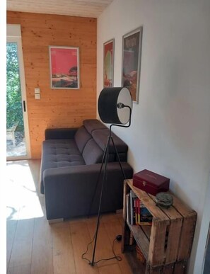 A room with a wooden wall, a grey sofa, a floor lamp, and a bookshelf with books and a red box.