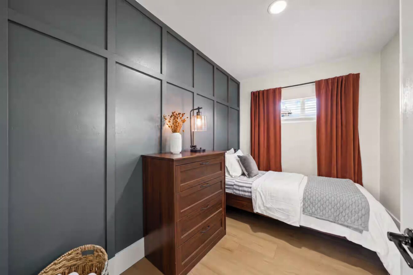 Ein Schlafzimmer mit Bett, Kommode, Wandlampe und einem Fenster mit Vorhängen.