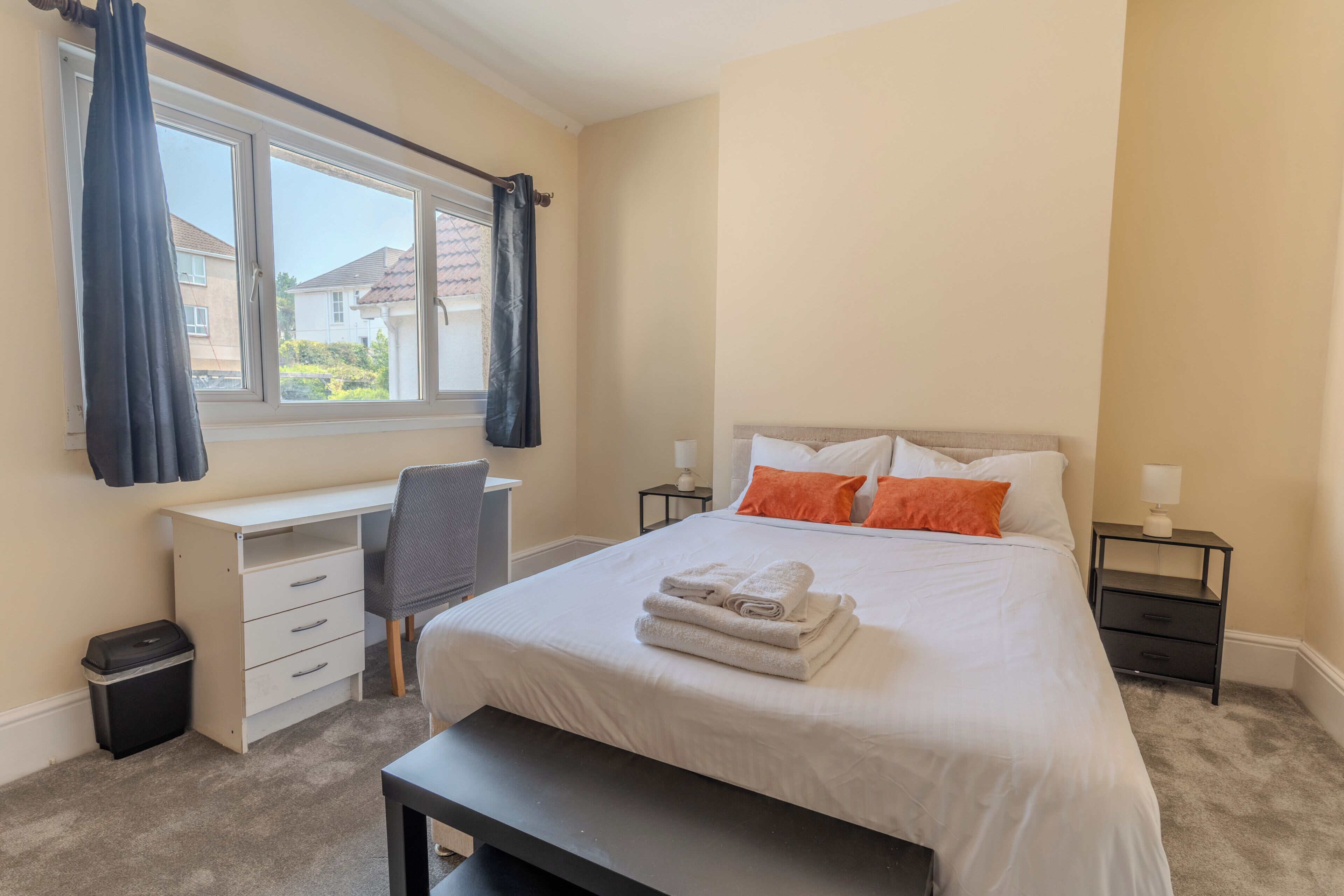 A bedroom with a bed, a desk, a chair, a bedside table, and a window with curtains.