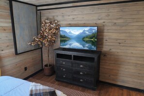 Ein Schlafzimmer mit einem Bett, einer Kommode, einem Fernseher an einer Holzwand und einer Zimmerpflanze.