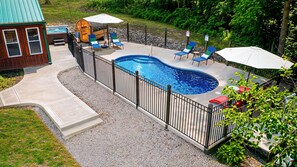 A swimming pool with a curved edge, surrounded by a black metal fence. There are lounge chairs and umbrellas around the pool. - Hocking Hills, Pool, Hottub & Sauna (Logan)