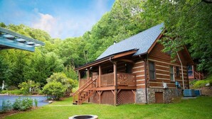 A log cabin with a porch, a stone foundation, and a small pond in the yard.