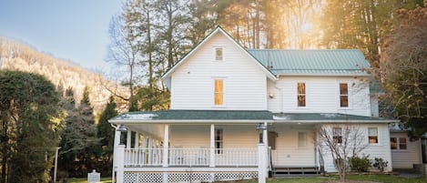 A two-story white house with a green roof, a covered porch, and a stone foundation.