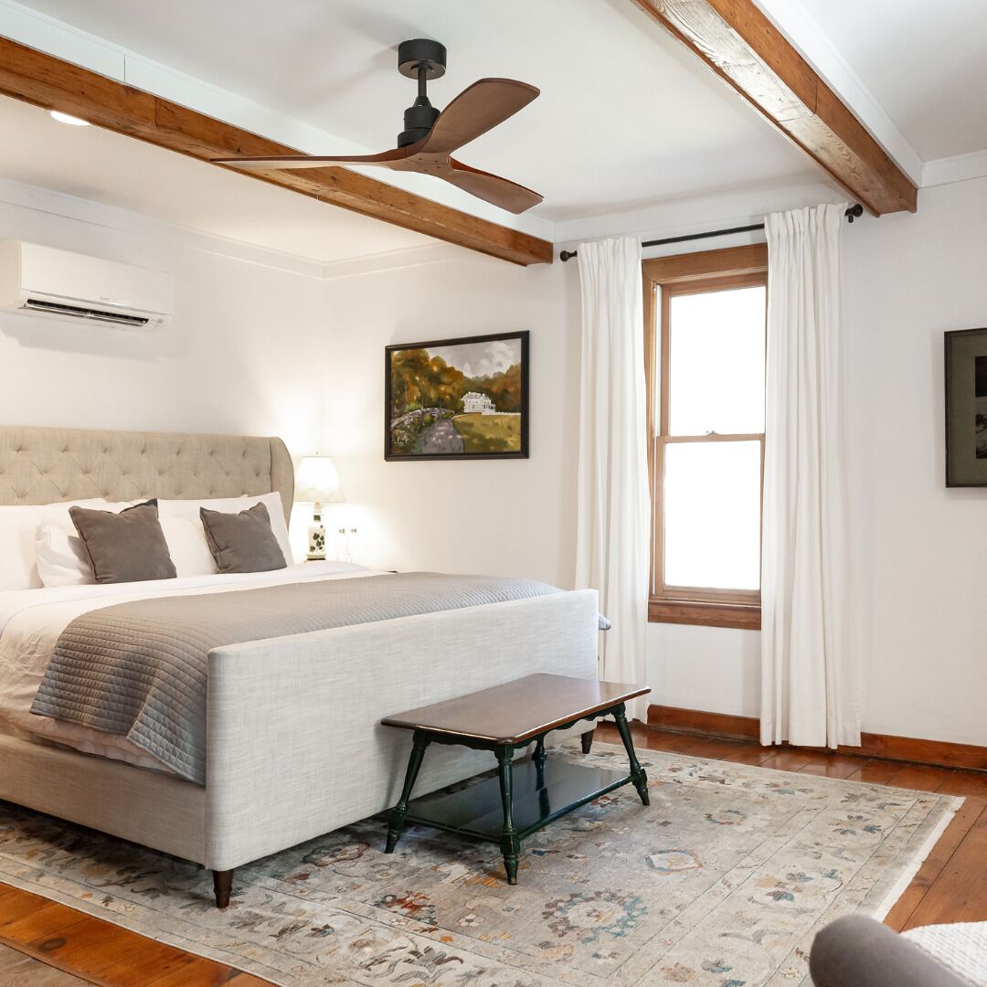 Un dormitorio con cama, un banco, ventilador de techo, ventana con cortinas y un cuadro en la pared.