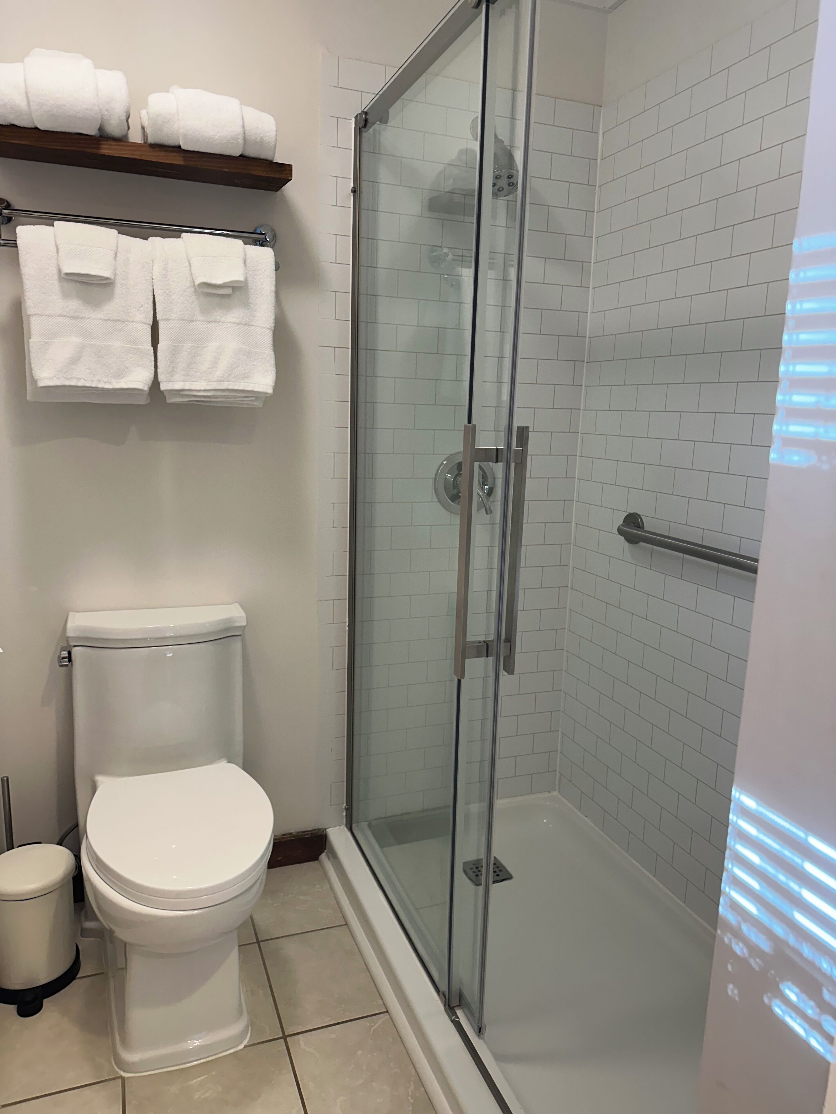 A bathroom with a glass-enclosed shower, a toilet, and a shelf with folded towels.