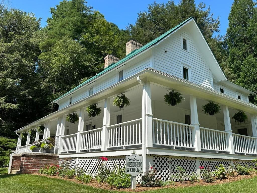 A white two-story house with a porch, a sign for guest registration, and a brick foundation.