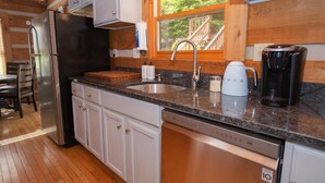 A kitchen with a stainless steel refrigerator, a dishwasher, a coffee maker, and a window with a view of a wooden deck.