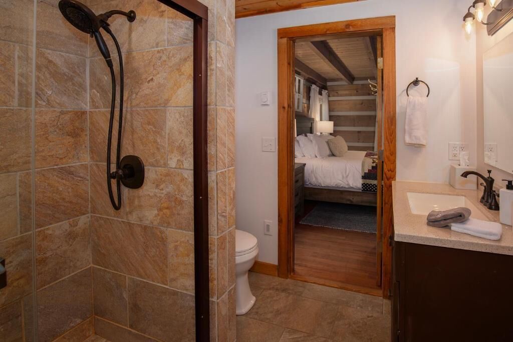 Une salle de bain avec douche, toilettes et une porte en bois donnant sur une chambre à coucher équipée d’un lit et d’une table de chevet.