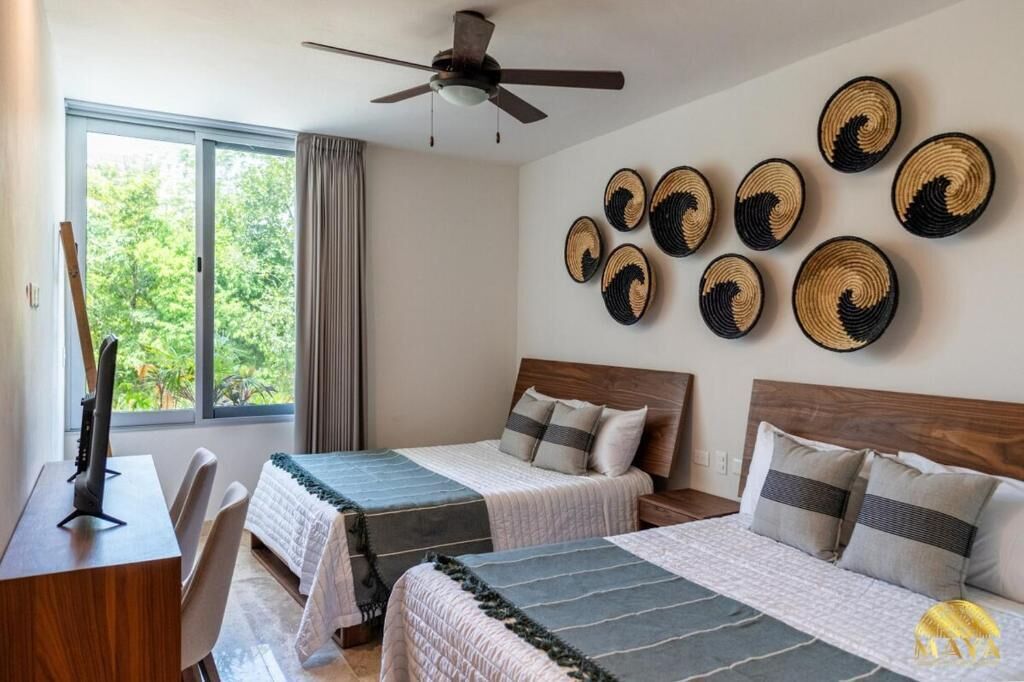 Quarto de hotel com duas camas, uma escrivaninha, ventoinha no teto e vista para o verde lá fora.