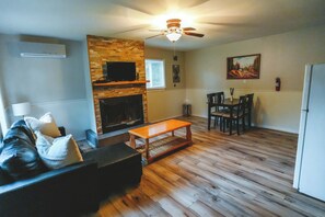 A living room with a fireplace, a ceiling fan, a dining area with chairs, and a television mounted on a stone wall. - Falcon Cottage - 2 Bedroom - Family Friendly (Trent Lakes)