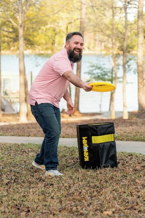 A person holding a yellow frisbee, standing next to a black and yellow bag on a grassy area with trees in the background.