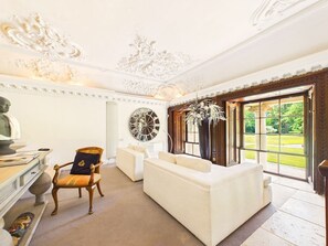 Una habitación con un sofá blanco, una silla de madera, un ventanal con vistas a zonas verdes y un techo decorativo.