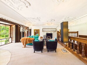 Een ruime kamer met een open haard, twee fauteuils, een bank en uitzicht op groen.