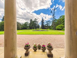 A view through columns to a well-manicured lawn with a fountain, surrounded by trees.