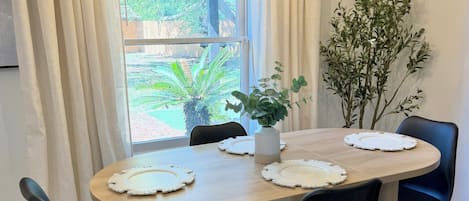 En matplats med ett runt bord, svarta stolar, en vas med växter och ett fönster med gardiner.