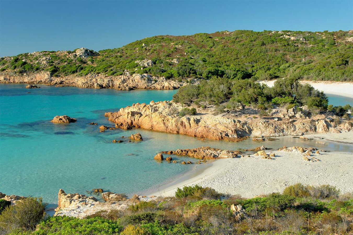 Paesaggio costiero con terreno roccioso, acque cristalline turchesi e una spiaggia sabbiosa.