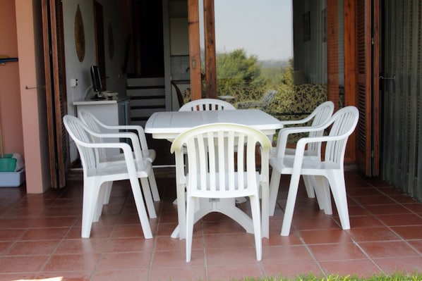 An outdoor patio with a white table and chairs, a tiled floor, and a view of greenery.
