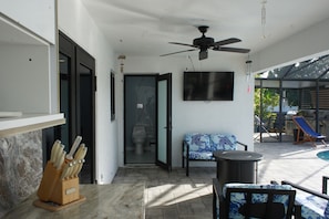A sunlit patio area with a ceiling fan, a television mounted on the wall, and a seating area with a blue patterned cushion. - Our Serenity Cove with a private pool in Cape Coral. (Cape Coral)