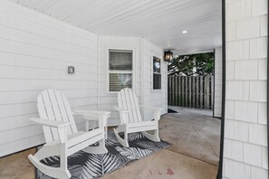 A covered patio area with two Adirondack chairs and a table, a wooden fence in the background, and a house with white siding. - North End 1BR/1BA Unit 3 - Walk to Beach & Boardwalk (Ocean City)